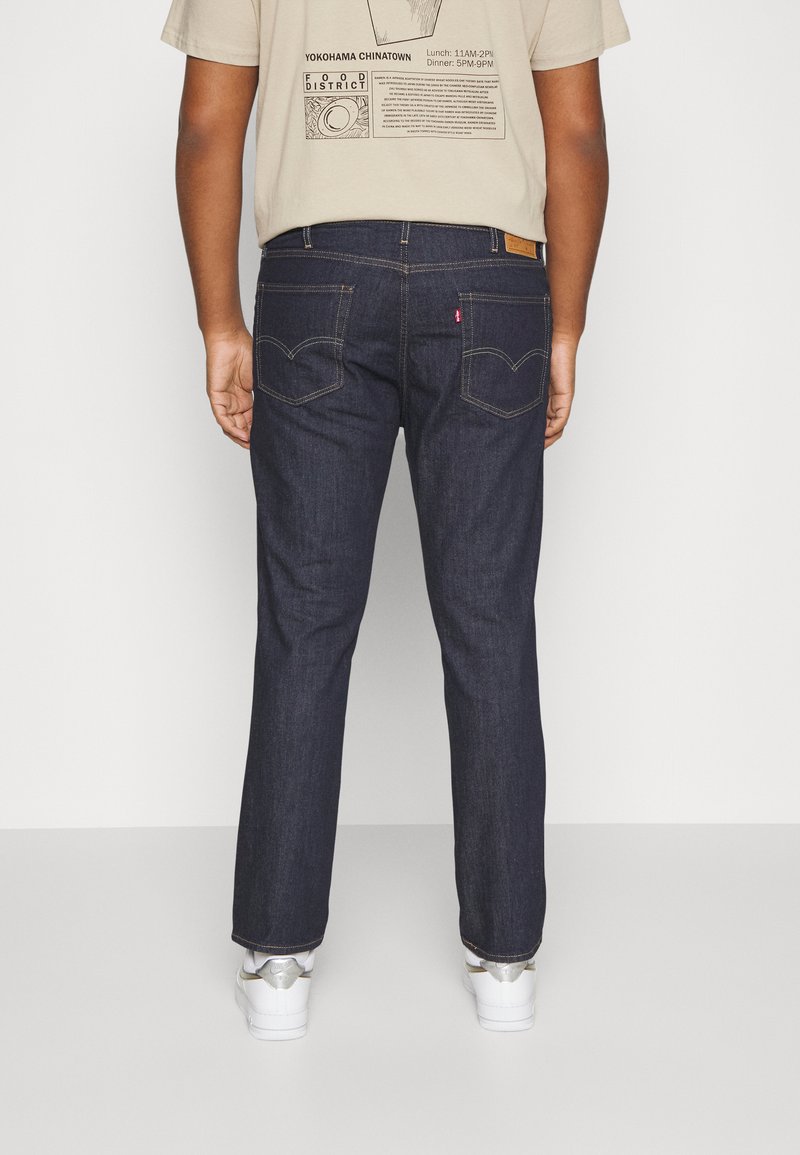 Jean en denim bleu foncé avec une coupe droite, poches arrière visibles et détails de couture subtils, associés à un T-shirt graphique beige.