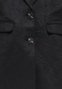 Cappotto in misto lana nero con una texture liscia, caratterizzato da due tasche con patta e due bottoni con cuciture a contrasto chiaro.