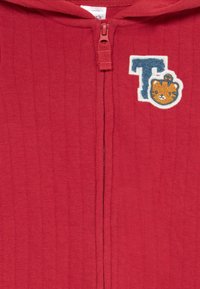 Sudadera roja con cremallera y textura acanalada, que presenta un parche bordado de un gato de dibujos animados al lado de la letra "T".