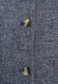 Υφασμα τζιν σε μπλε απόχρωση με υφή, που διαθέτει ένα στρογγυλό ανοιχτόχρωμο κουμπί με τέσσερις τρύπες.
