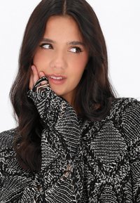 Pulover tricotat cu model alb și negru, cu mâneci lungi, având un material texturat și manșete fără degete, creând un aspect stratificat.