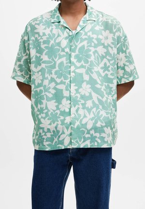 Mann trägt kurzärmeliges Hemd in Grün mit weißem Blumenmuster und dunkelblaue Jeans vor neutralem Hintergrund.