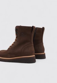 Botas de cuero marrón con una textura suave, cordones en la parte delantera, cremallera lateral y costuras en contraste en color crema en la suela. Diseño duradero y robusto.