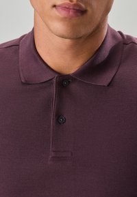 Polo bordeaux en tissu texturé, avec un col classique et des boutons noirs. Vue en gros plan mettant en valeur l'encolure et les coutures.