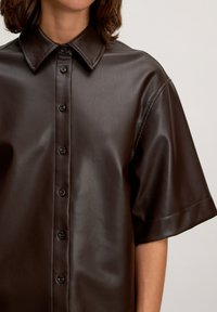 Braunes Fauxlederhemd mit spitzem Kragen, kurzen Ärmeln und vorderem Knopfverschluss. Glatte Textur mit einem klaren, modernen Design.