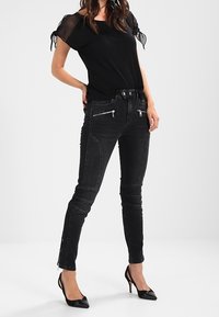 Jeans de denim negro con bolsillos con cremallera, corte ajustado y textura suave. Combinados con una blusa negra con mangas transparentes y detalles de lazo.