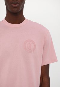 Camiseta de algodón rosa con cuello redondo. Presenta un logo sutilmente grabado en el pecho en un color a juego, con una textura suave.
