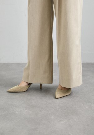 Des chaussures à talons beiges avec un bout pointu, présentant une texture pailletée. Portées avec un pantalon ample de couleur claire. Fond de surface plane.
