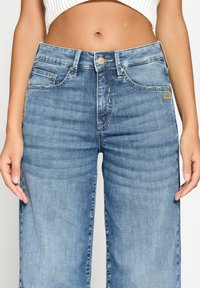 Střední část osoby oblečené v jasně modrých džínových kalhotách s vysokým pasem a bílém zkráceném pleteném topu, stojící s uvolněnýma rukama podél těla.