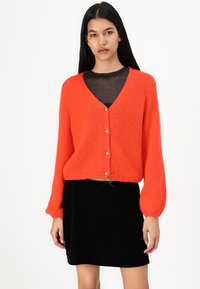 Cardigan orange vif avec un col en V, texture tricot épaisse et fermeture à trois boutons. Associé à une jupe en velours noir.