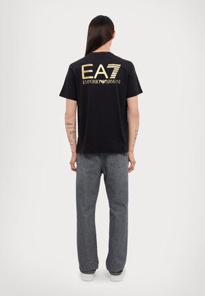 Personne aux longs cheveux foncés, de dos, portant un t-shirt noir avec le texte doré "EA7 Emporio Armani" et un jean gris.