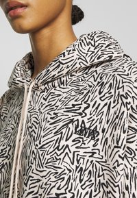 Närbild av en person som bär en beige hoodie med svarta abstrakta zickzackmönster och en liten broderad logga på bröstet.