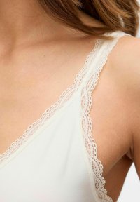 Camisola bianca con delicata bordatura in pizzo a scallop lungo le spalline, caratterizzata da una texture liscia e un design aderente. Vista ravvicinata della zona delle spalle.