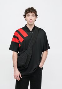 Čierna košeľa s krátkym rukávom s červenými diagonálnymi pruhmi, výstrih do V a matná textúra. Modelka nosí čiernu crossbody tašku s pruhmi.