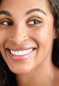 Gros plan d'un visage souriant avec une peau brun moyen, des lèvres brillantes et des yeux marron. La peau a une texture lisse avec des taches de rousseur subtiles.