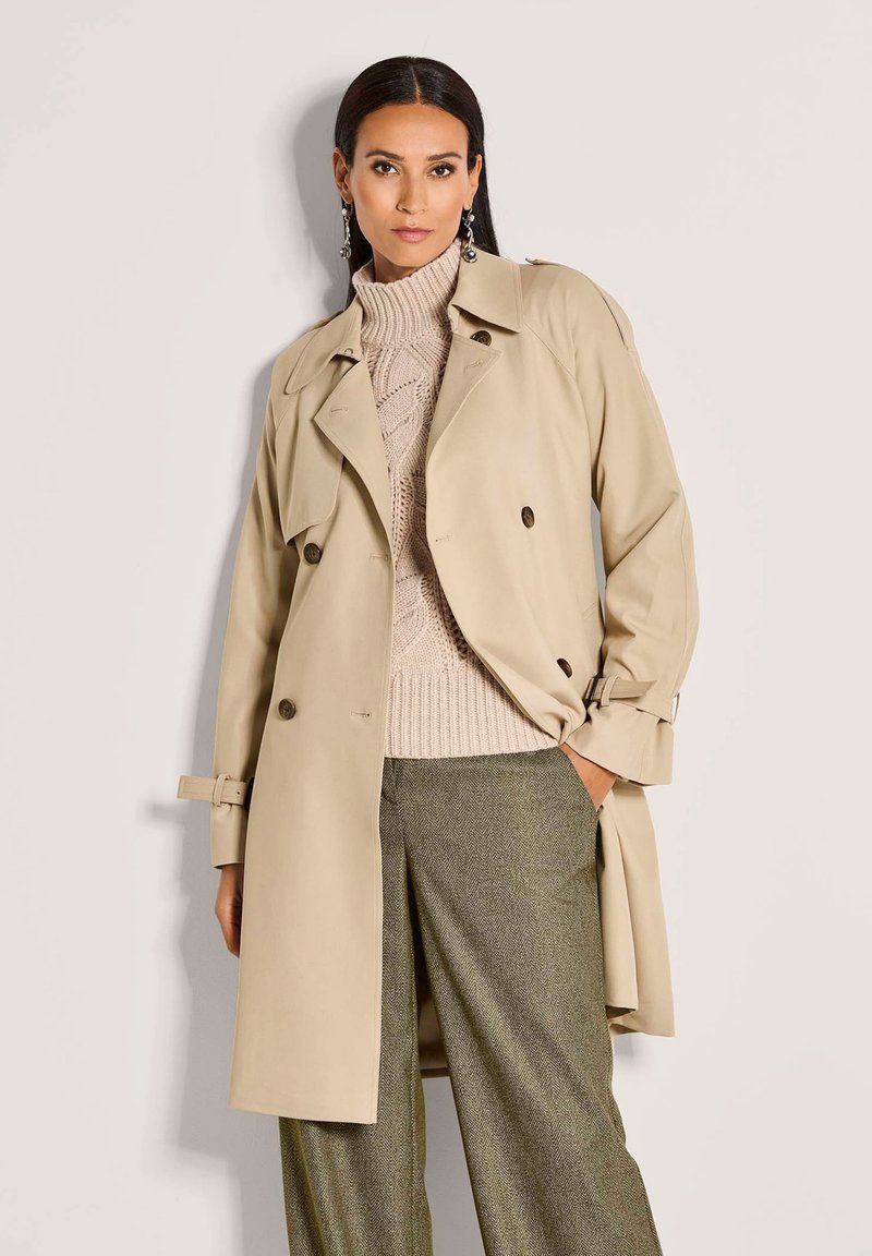 Vrouw in een beige trenchcoat over een kabelgebreide coltrui en grijze wijde broek, staande tegen een effen achtergrond.