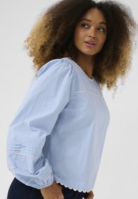 Blusa di cotone azzurro chiaro con maniche a sbuffo, orlo ondulato e cuciture dettagliate lungo il collo e i polsini delle maniche.
