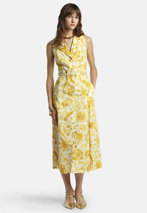 Nő zsebébe tett kézzel áll, ujjatlan sárga, virágos, gombos és öves midi ruhát visel, arany magassarkúval és karkötővel párosítva.