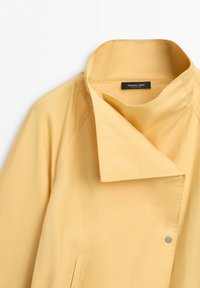 Hellgelber Mantel mit hohem Kragen, asymmetrischer Frontklappe und sichtbarem Druckknopf, mit Massimo Dutti Etikett innen am Kragen.