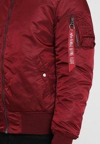 Rode bomberjack met een gladde, glanzende buitenkant, geribbelde manchetten, zijzakken en een verstelbare ritszak met een label waarop staat "REMOVE BEFORE FLIGHT".