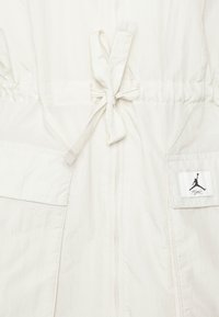 Veste blanche légère avec une taille élastique, un cordon à nouer, une fermeture éclair et deux poches avant ornées d'un écusson logo noir.