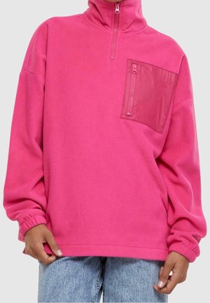 Persoon draagt een felroze fleece trui met halve rits en een met rits afsluitbare borstzak, gecombineerd met lichtblauwe spijkerbroek tegen een effen achtergrond.