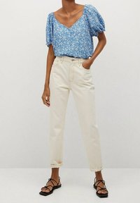 Blouse florale bleu clair avec des manches bouffantes, associée à un pantalon crème taille haute et des sandales noires à lanières. Tenue décontractée.
