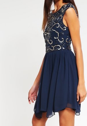 Femme portant une robe bleu marine sans manches avec un corsage ajusté orné de sequins et une jupe fluide en chiffon, debout devant un fond uni.