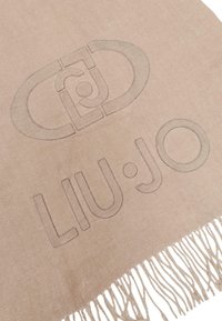 Bufanda beige con un sutil diseño de logo en relieve y bordes con flecos, hecha de un material suave para un acabado texturizado.