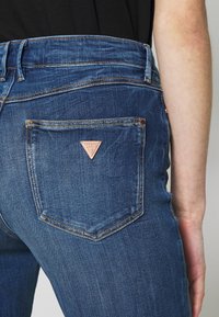 Mörkblå denimjeans med en figursydd design, som har en triangelformad patch på bakfickan och kontrasterande sömnadsdetaljer.