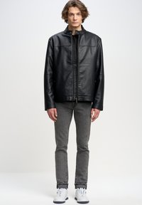 Chaqueta de cuero negra con cuello alto y cierre de cremallera. Combinada con jeans ajustados grises y zapatillas blancas. Textura suave, diseño casual.
