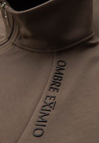 Sudadera con cremallera en color marrón que presenta un cuello alto, una textura suave y texto negro en relieve que dice "OMBRE EXIMIO" a lo largo de la costura lateral.