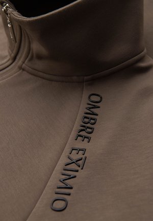 Pullover marrone con zip, caratterizzato da un colletto alto, una texture morbida e testo nero in rilievo che recita "OMBRE EXIMIO" lungo la cucitura laterale.