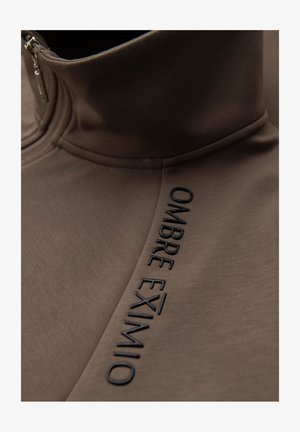 Pullover marrone con zip, caratterizzato da un colletto alto, una texture morbida e testo nero in rilievo che recita "OMBRE EXIMIO" lungo la cucitura laterale.