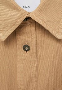 Camicia button-up beige con colletto a punta, tessuto testurizzato e dettaglio in metallo al collo. Etichetta del brand visibile all'interno del colletto.