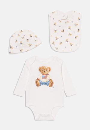 Fehér bébi body, amelyen egy plüssmackó grafika látható farmernadrágban és pólóban, amerikai zászló mintával. Kiegészül egy mintás, illő sapkával és bíbimmel.