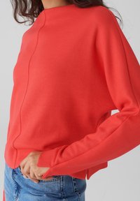 Pull rouge à col montant, à manches longues et à coupe décontractée. Présente des fentes latérales et une surface lisse et texturée. Associé à un jean.