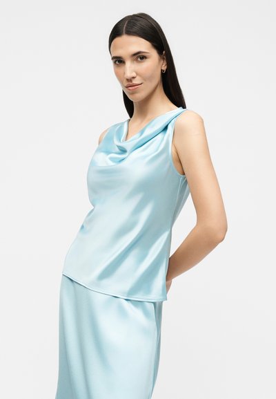 Γυναίκα με μακριά σκούρα μαλλιά, που φοράει ένα αμάνικο ανοιχτό μπλε σατέν τοπ και φούστα που ταιριάζει, στέκεται μπροστά σε ένα απλό λευκό φόντο.