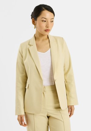 Femme aux cheveux foncés portant un blazer crème et un pantalon assorti, un haut blanc à col en V et des boucles d'oreilles créoles dorées, regardant vers sa gauche.