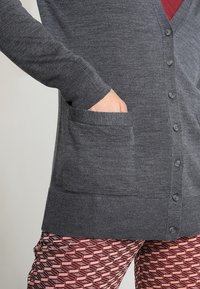 Grå stickad cardigan med V-hals, framknappar och två sidofickor, kombinerad med mönstrade rosa byxor med geometriska mönster.