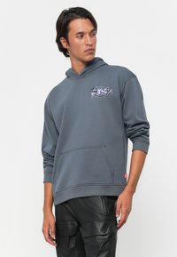 Noor mees, kes kannab sinist kapuutsi sweatshirt'i, millel on ees tasku, ja musti nahast cargo-pükse, vaatab kõrvale valge tausta ees.