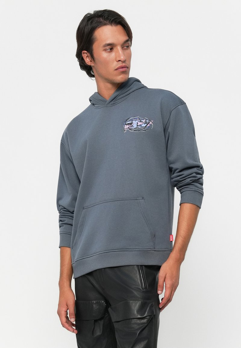 Joven con un sudadera con capucha azul y bolsillo frontal, y pantalones cargo de cuero negro, mirando hacia un lado frente a un fondo blanco.