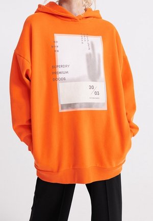 Przewymiarowana jasnopomarańczowa bluza z kapturem z dużym białym prostokątnym nadrukiem z napisem "SUPERDRY PREMIUM GOODS" oraz "20/03 ESTABLISHED" na przodzie.