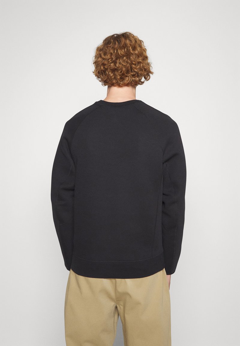 Sweatshirt noir avec un col rond côtelé et des manches raglan. Texture lisse, sans motifs visibles, associé à un pantalon beige.