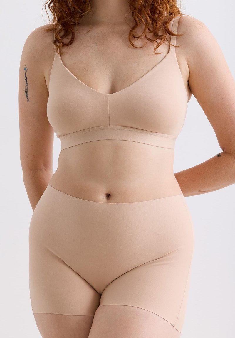 Frau mit roten lockigen Haaren, die einen nahtlosen BH in Nude sowie Unterwäsche im Boyshort-Stil trägt und Tattoos auf den Oberarmen zeigt, vor einem schlichten Hintergrund.