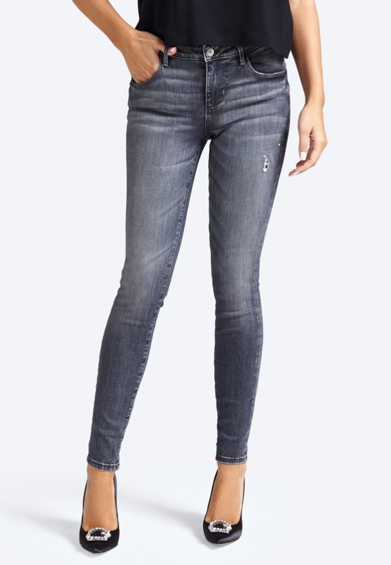 Mujer que lleva jeans ajustados grises con un ligero desgaste en el muslo, blusa negra y tacones negros con hebillas decorativas, posando con una mano en el bolsillo.