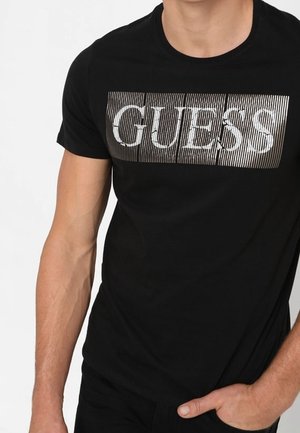Mann trägt ein schwarzes Kurzarmshirt mit einem weißen, gestreiften rechteckigen Logo mit der Aufschrift "GUESS" auf der Brust.
