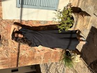 Jeune femme aux longs cheveux foncés portant une robe noire et des talons bleus, debout contre un mur en pierre rustique ensoleillé avec des plantes en pot.