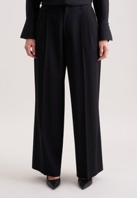 Zwarte wijde broek van gladde stof met een voorplooien. Gekleed met zwarte puntige schoenen, met een op maat gemaakte tailleband detail.