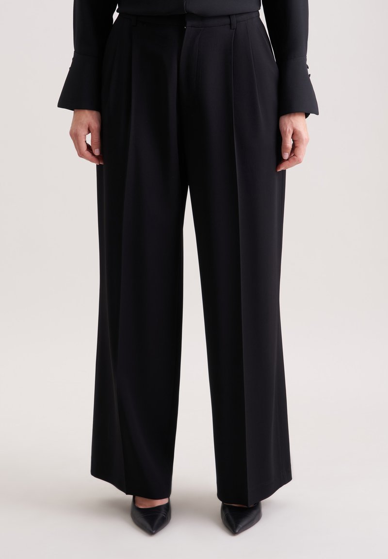 Zwarte wijde broek van gladde stof met een voorplooien. Gekleed met zwarte puntige schoenen, met een op maat gemaakte tailleband detail.